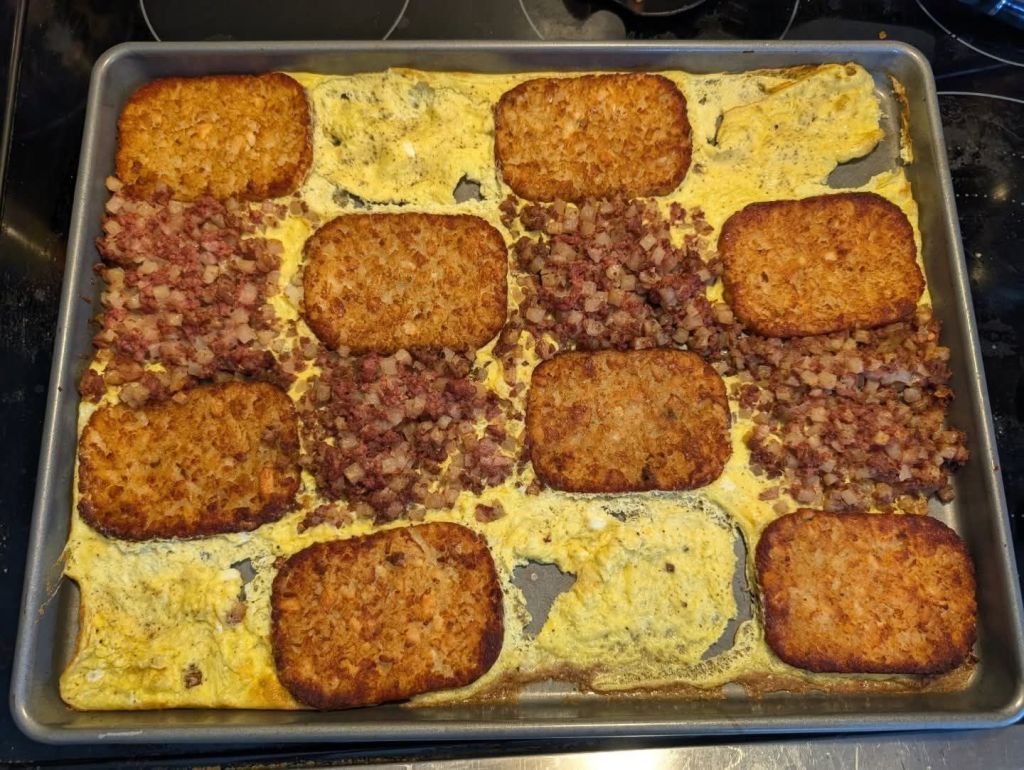 A baking sheet filled with golden-brown hash brown patties arranged in a grid, with sections of scrambled eggs and corned beef hash interspersed between them.