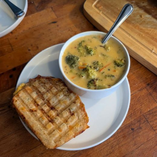 Golden roasted broccoli cheddar soup with pops of green and orange (from carrots) in a small white bowl with a spoon sitting beside a panini-grilled ham & cheese sandwich with cross-hatch lines on a white plate.  It's all on a wooden counter top, beside a wooden cutting board,
