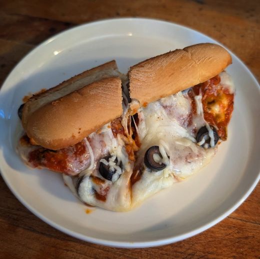Oven-toasted meatball sub sliced in half on a small round white plate, served atop a warm wooden surface. The sub roll is stuffed with frozen meatballs, rich tomato sauce, black olive slices, and a gooey blend of melted provolone and mozzarella cheese, lightly browned from broiling. The filling spills slightly from the cut edges, showcasing a rustic, hearty presentation... but also calling to the great eldritch god Cthulhu climbing out of the sea.