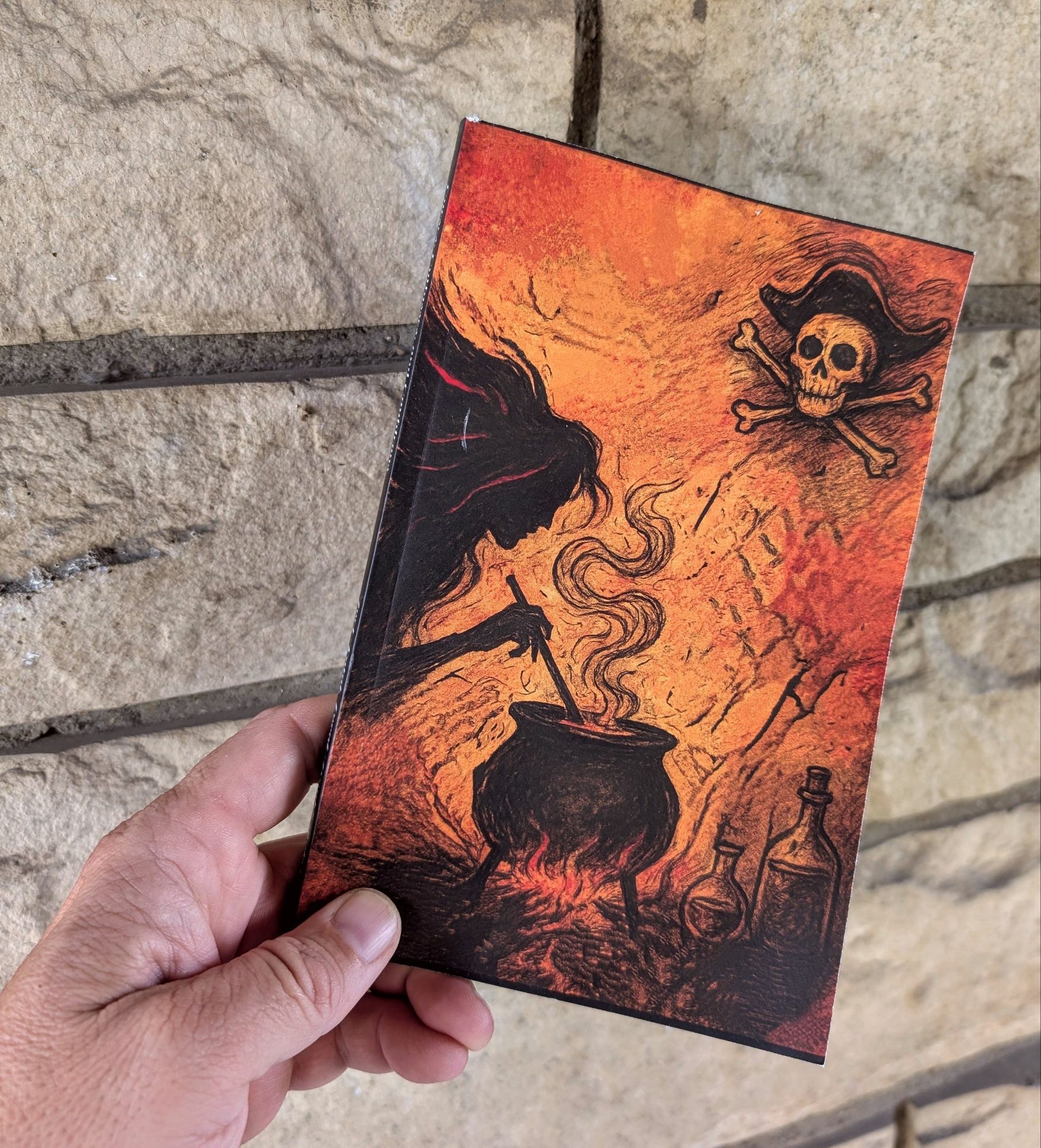 A hand holding a book with an illustration of a witch stirring a cauldron against an orange background, featuring a skull and crossbones in the upper corner.