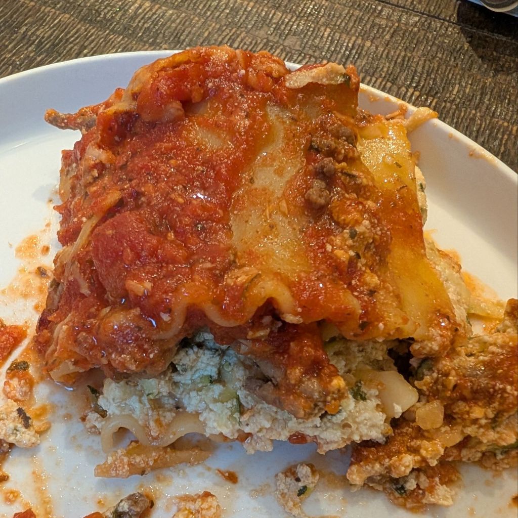 A serving of lasagna on a white plate, showcasing layers of noodles, marinara sauce, ricotta cheese, and ground beef.