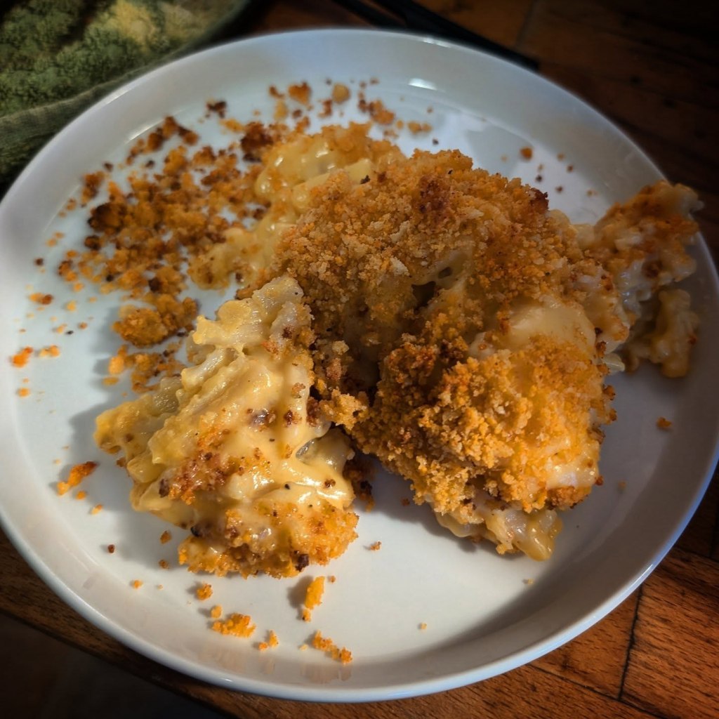 Creamy baked mac and cheese on a white plate, topped with golden breadcrumbs. The dish is served with crumbled crumbs scattered around.