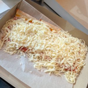 A cut from Beto's | A slice of cheesy pizza with a thick layer of unmelted shredded cheese in a takeout box, resting on parchment paper.