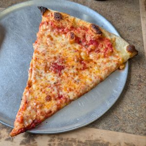 A slice from Slice on Broadway | A slice of cheese pizza on a metal tray, featuring a golden-brown crust and melted cheese with a slightly crispy edge.