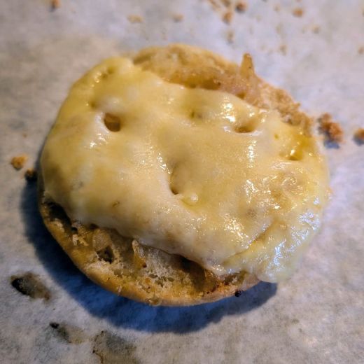 A close-up of a toasted English muffin topped with caramelized onions & melted Swiss cheese on a parchment paper background. 🍞🧈🧅🧀