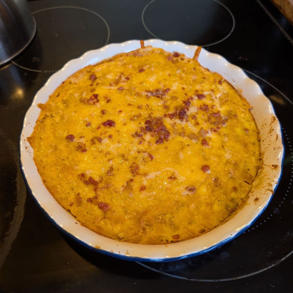 A baked casserole with a golden cheesy topping, garnished with bits of bacon, in a blue-trimmed white pie dish on a stovetop.