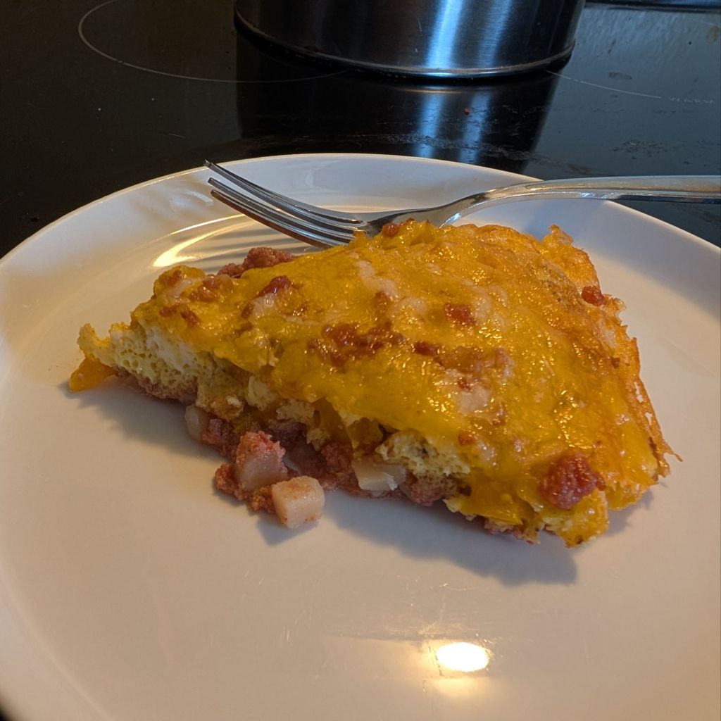 A slice of cheesy corned beef hash breakfast bake, with visible ham and potato on a white plate, accompanied by a fork.