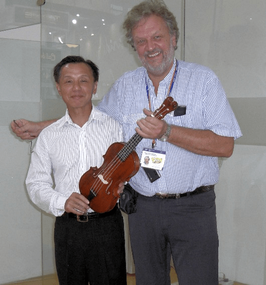 Jerker Antoni and Colleague with Violin-Shaped Ukulele: A candid photo of two men smiling together indoors, with the man on the right—Jerker Antoni—wearing a striped button-down shirt and holding a small, brown violin-shaped instrument or ukulele, with a conference badge hanging from a lanyard.