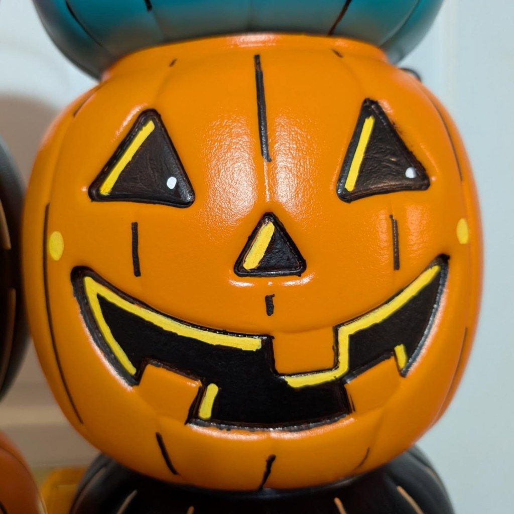 A close-up shot of an orange plastic jack-o'-lantern bucket with painted black triangular eyes, a small triangular nose, and a wide grinning mouth, with the top of a teal bucket visible just above it.