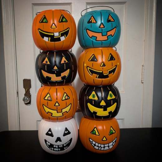 Two tall stacks of colorful plastic jack-o'-lantern trick-or-treat buckets standing on a dark surface in front of a white door. The left stack features orange, black, orange, and white buckets from top to bottom. The right stack features teal, orange, black, and orange buckets.
