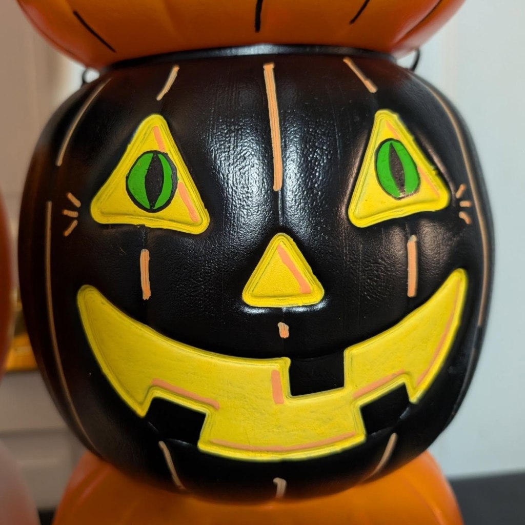 A close-up of a black plastic jack-o'-lantern bucket featuring bright green cat-like eyes with vertical pupils inside yellow triangles, a yellow nose, and a wide yellow-lined grin