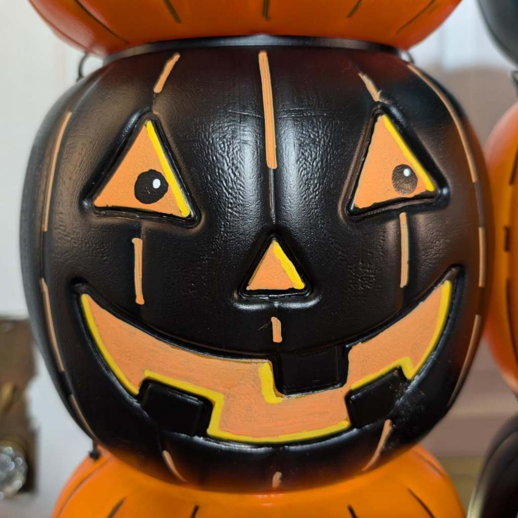 A close-up of a black plastic jack-o'-lantern bucket featuring painted orange triangular eyes with small black pupils, an orange nose, and an orange-accented smiling mouth