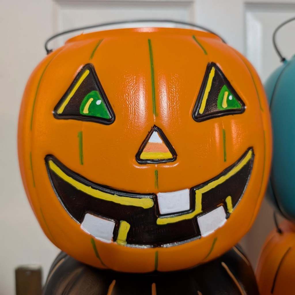 A close-up of an orange plastic jack-o'-lantern bucket featuring green cat-like eyes, a nose painted to look like a piece of candy corn, and a broad smile showing white teeth.