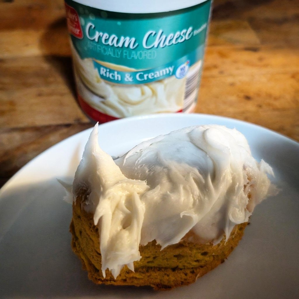 A close-up of a golden-brown baked Halloween treat on a white plate, thickly frosted with white cream cheese frosting, with a tub of Baker's Corner cream cheese frosting sitting blurred in the background.