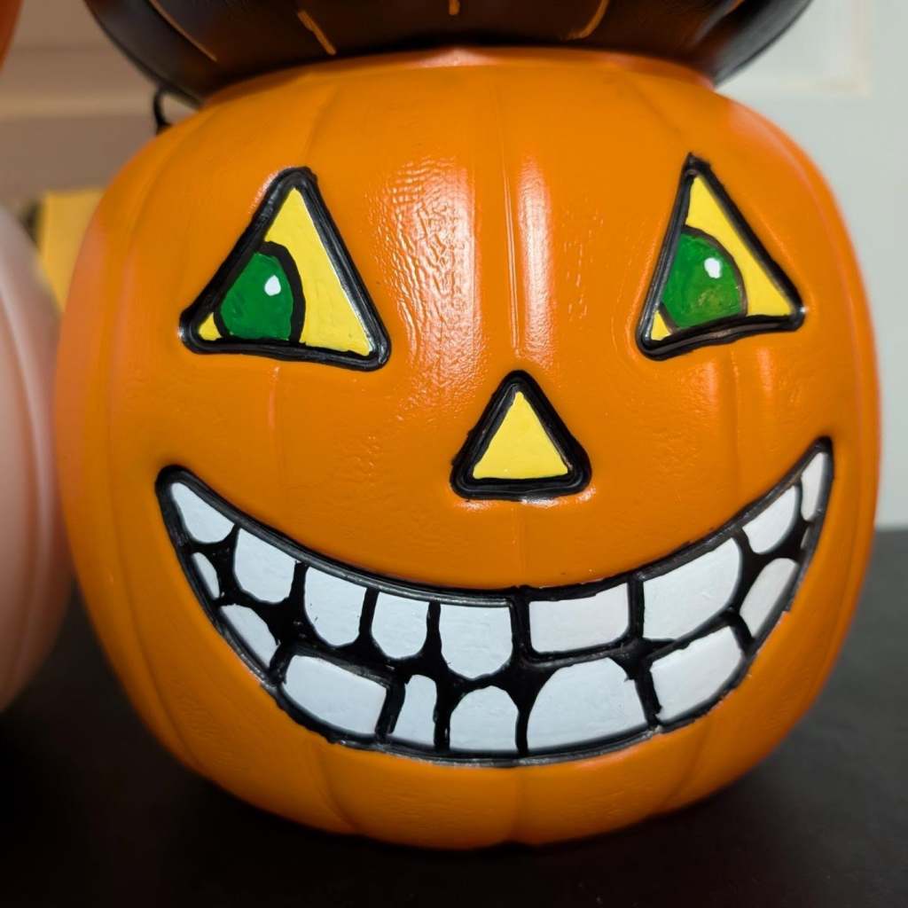 A close-up of an orange plastic jack-o'-lantern bucket featuring green eyes inside yellow triangles and a wide, mischievous grin filled with many white teeth.