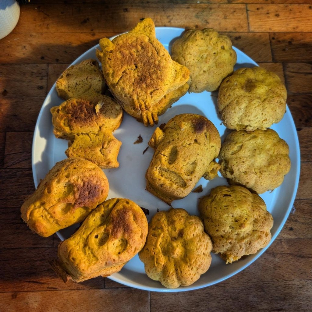 A white plate filled with an assortment of freshly baked, golden-brown Halloween-themed cakes shaped like skulls, pumpkins, a bat, cats, and Frankenstein's monster.