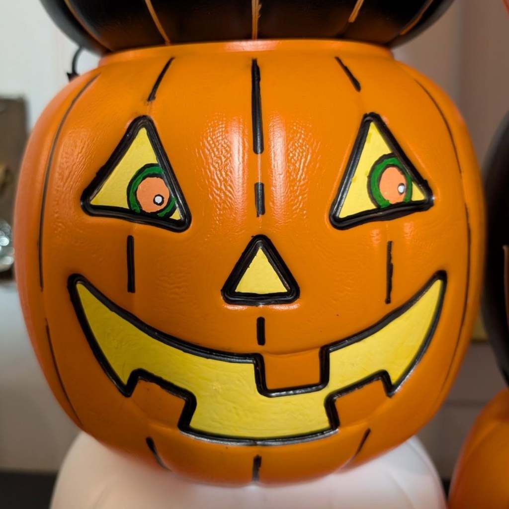 A close-up of an orange plastic jack-o'-lantern bucket featuring triangular eyes with round, surprised-looking orange-and-green pupils, a small nose, and a classic carved grin.