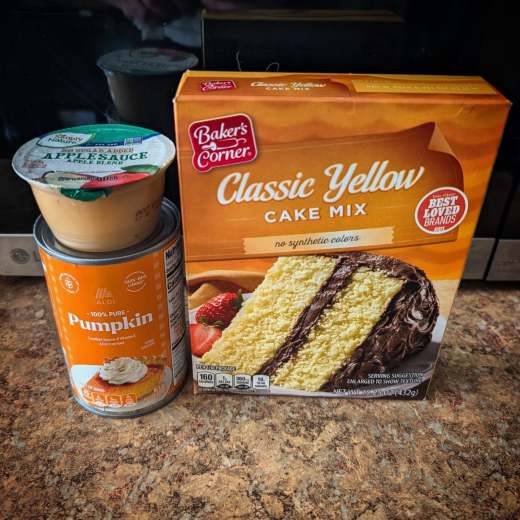 A countertop view of the baking ingredients used, featuring a box of Baker's Corner Classic Yellow cake mix, a can of 100% pure pumpkin, and a cup of applesauce.