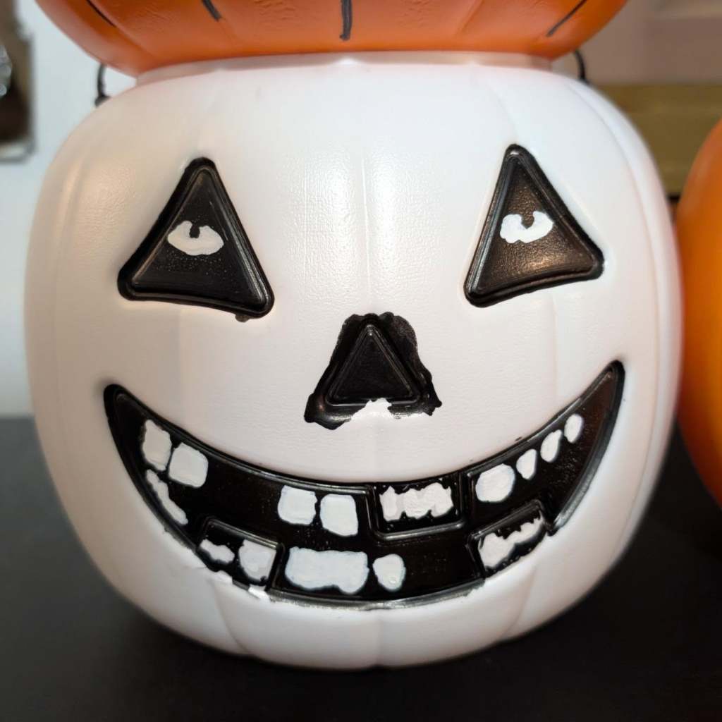 A close-up of a white plastic jack-o'-lantern bucket with black triangular eyes containing white highlights, a black nose, and a wide black-and-white toothy smile. (An amalgamation of a classic 🎃 & the Misfits' FIEND or Crimson Ghost logo.)
