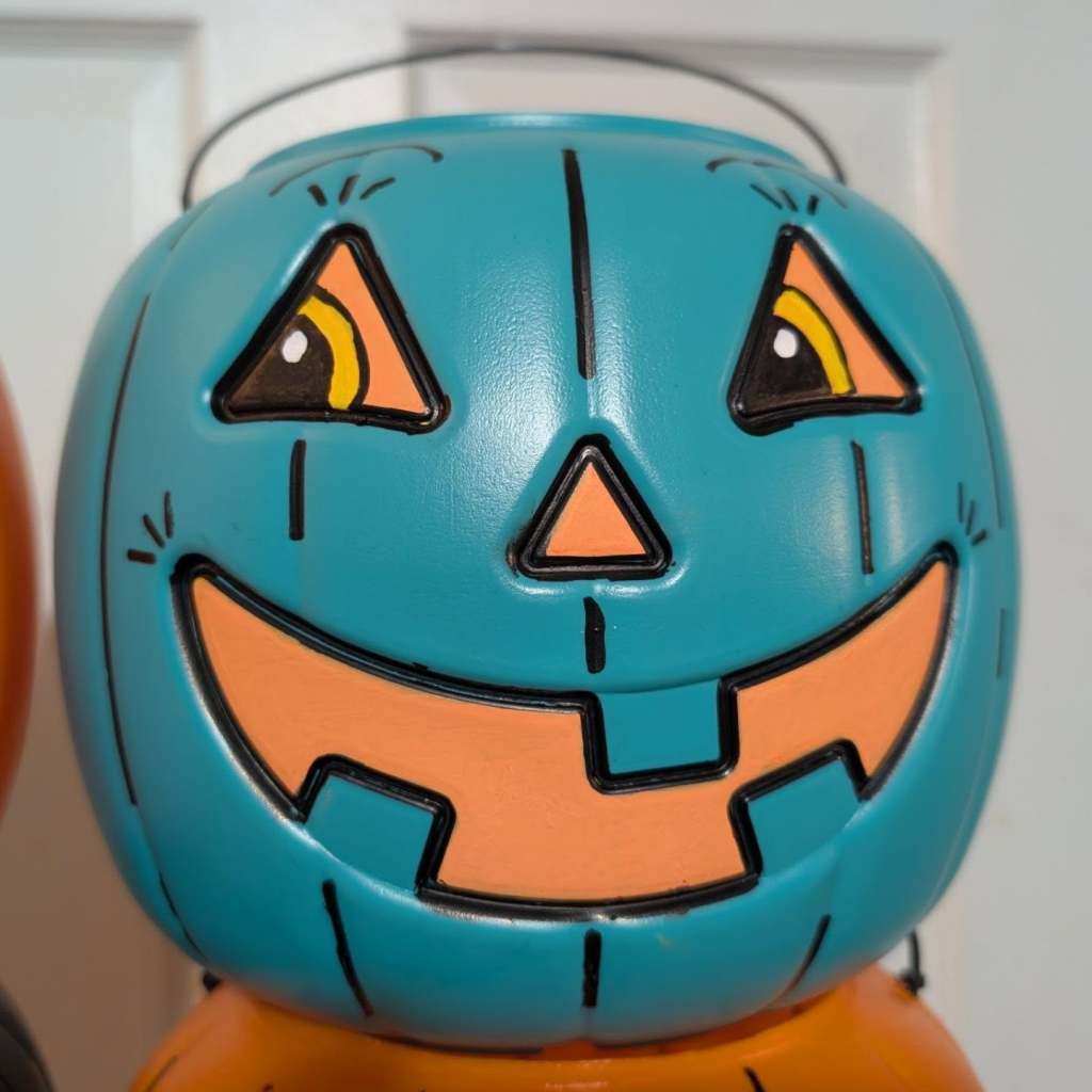 A close-up of a teal blue plastic jack-o'-lantern bucket featuring stylized orange-and-black eyes with short painted eyebrows and eyelashes, an orange nose, and a wide orange-lined grin. (For the Teal Pumpkin Project & Food Allergy Awareness!)