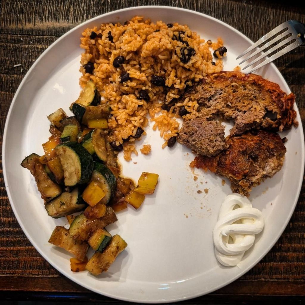 Sauteed zucchini & bell pepper, Spanish rice with black beans, and taco-night meatloaf on a plate next to a dollop of sour cream.