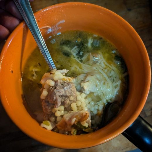 Overhead view of a bright orange soup bowl with a black handle, held in one hand. A spoon lifts a meatball resting on acini di pepe, with shredded chicken, melted cheese, wilted spinach, chopped carrots, and chopped celery floating in a golden broth.
